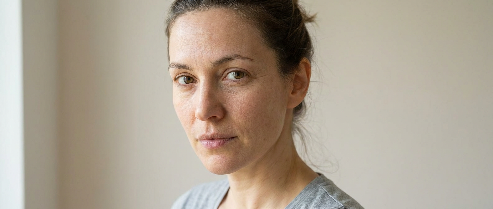 Portrait einer Frau vor hellem Hintergrund mit müder wirkender Haut, sichtbaren Hautfalten und ungleichmäßigem Hautbild, ohne Produkt.