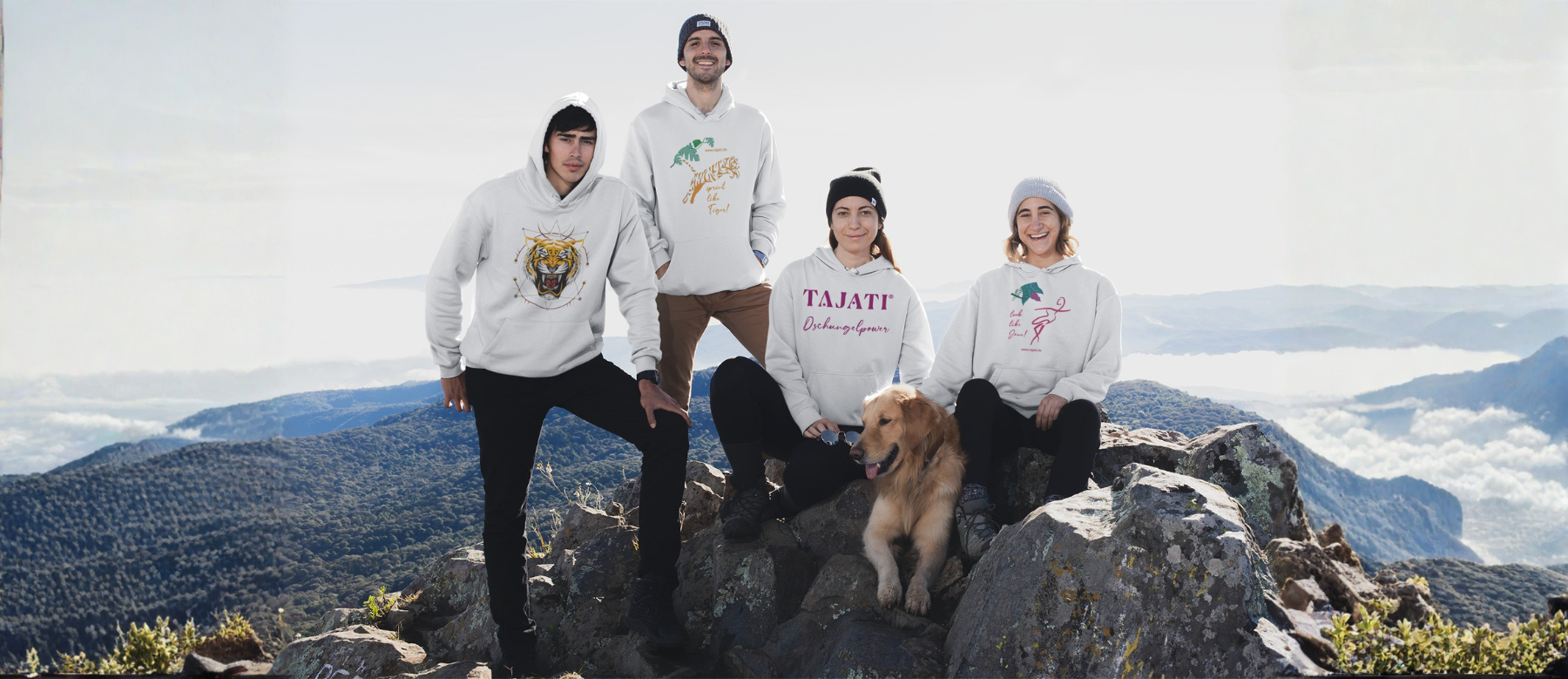 Banner der TAJATI® Kollektion: vier Personen in TAJATI-Hoodies und Mützen auf einem Berggipfel, daneben ein Hund. Textoverlay mit Button zur TAJATI® Kollektion für Bekleidung (Spreadshirt)